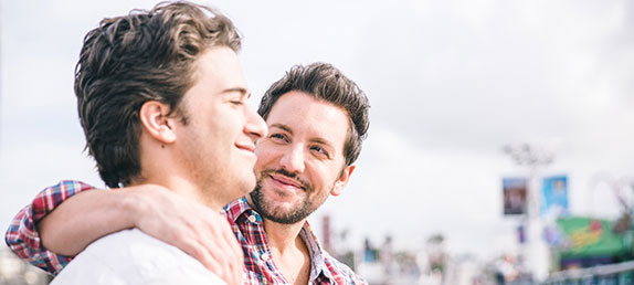 2 homosexual men sitting on a park bench with their arms around each other. Gay pride and gay marriage are a sinful choice.